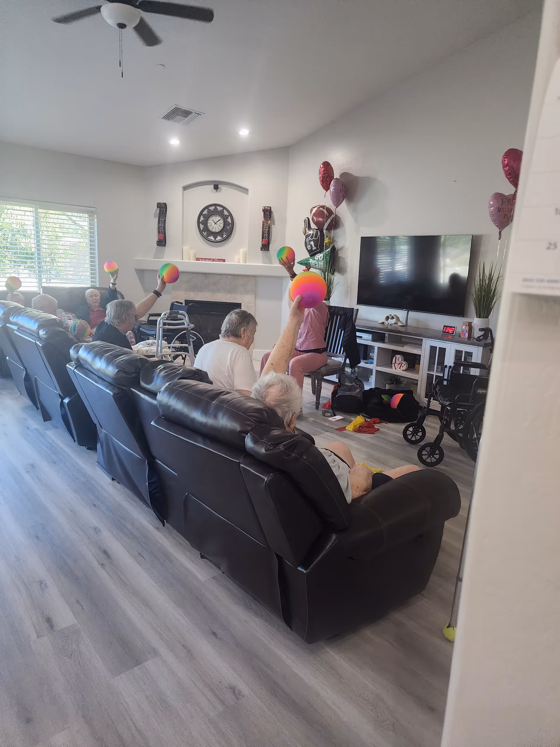 A group of elderly people seated on black leather recliners in a living room, participating in an activity where they hold up colorful balls. The room has a large window with blinds, a wall-mounted TV, a clock above a fireplace, and some balloons and decorations on the wall.