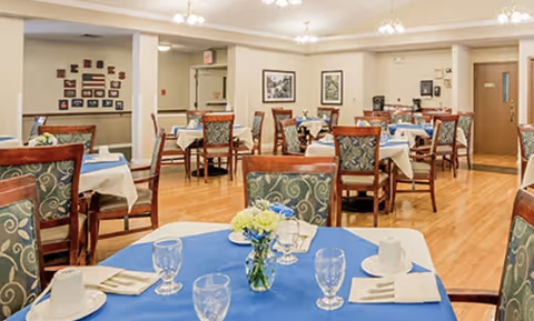 A dining room in a senior living facility with multiple tables covered in white and blue tablecloths. Each table is set with glasses, plates, napkins, and silverware. The chairs have patterned upholstery, and there are framed pictures on the walls. The room has wooden flooring and soft lighting from ceiling fixtures.