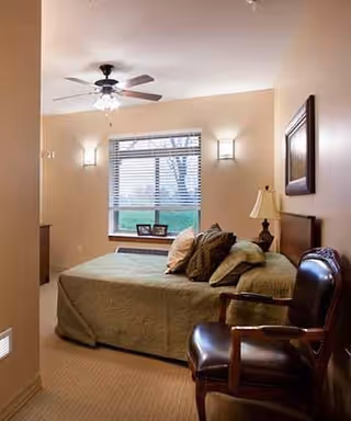 A cozy bedroom with a bed covered in a green quilt and multiple pillows. There is a wooden chair with leather upholstery in the foreground, a bedside table with a lamp, and a framed picture on the wall. A window with blinds lets in natural light, and two wall sconces provide additional lighting. A ceiling fan with lights is mounted on the ceiling.