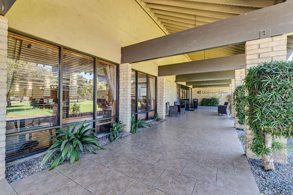 Covered outdoor walkway with tiled floor and large windows on one side showing an interior seating area. The walkway is supported by brick columns and wooden beams, with potted plants and chairs along the path. A sign on the far wall reads 'A Welcome Home'.