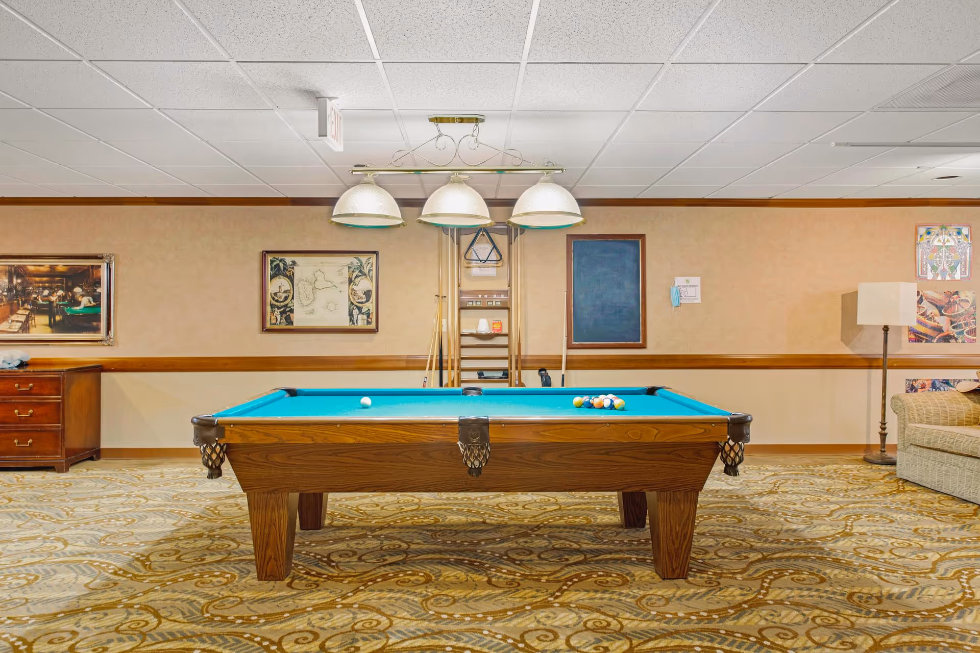 Interior view of a recreational room featuring a pool table with balls arranged for a game. The room has patterned carpet flooring, beige walls with framed pictures, a wooden cabinet on the left, a floor lamp and a sofa on the right. Overhead, three dome-shaped lights hang above the pool table.
