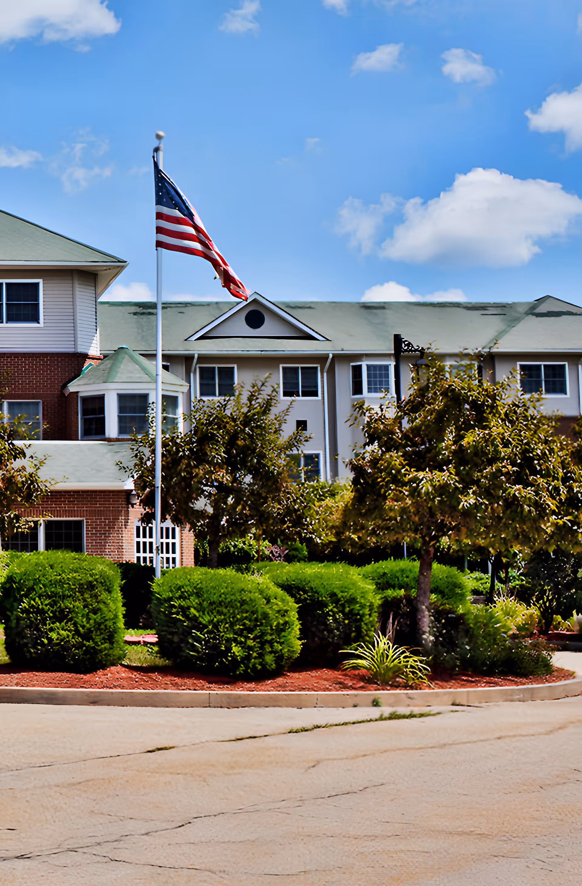 Exterior view of a senior living facility named Elan Gardens with a green roof, multiple windows, and a well-maintained garden with bushes and trees in front. An American flag is flying on a flagpole near the entrance under a blue sky with some clouds.