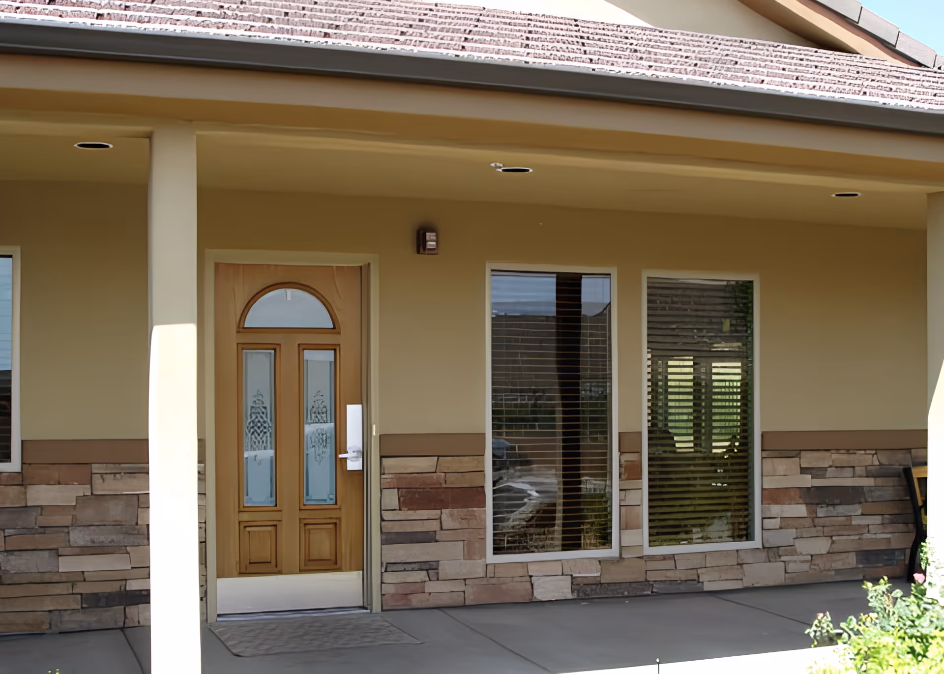 Exterior view of a building entrance with a wooden door featuring decorative glass panels and two large windows with blinds. The building has a beige facade with stone accents along the lower half and a covered porch area.