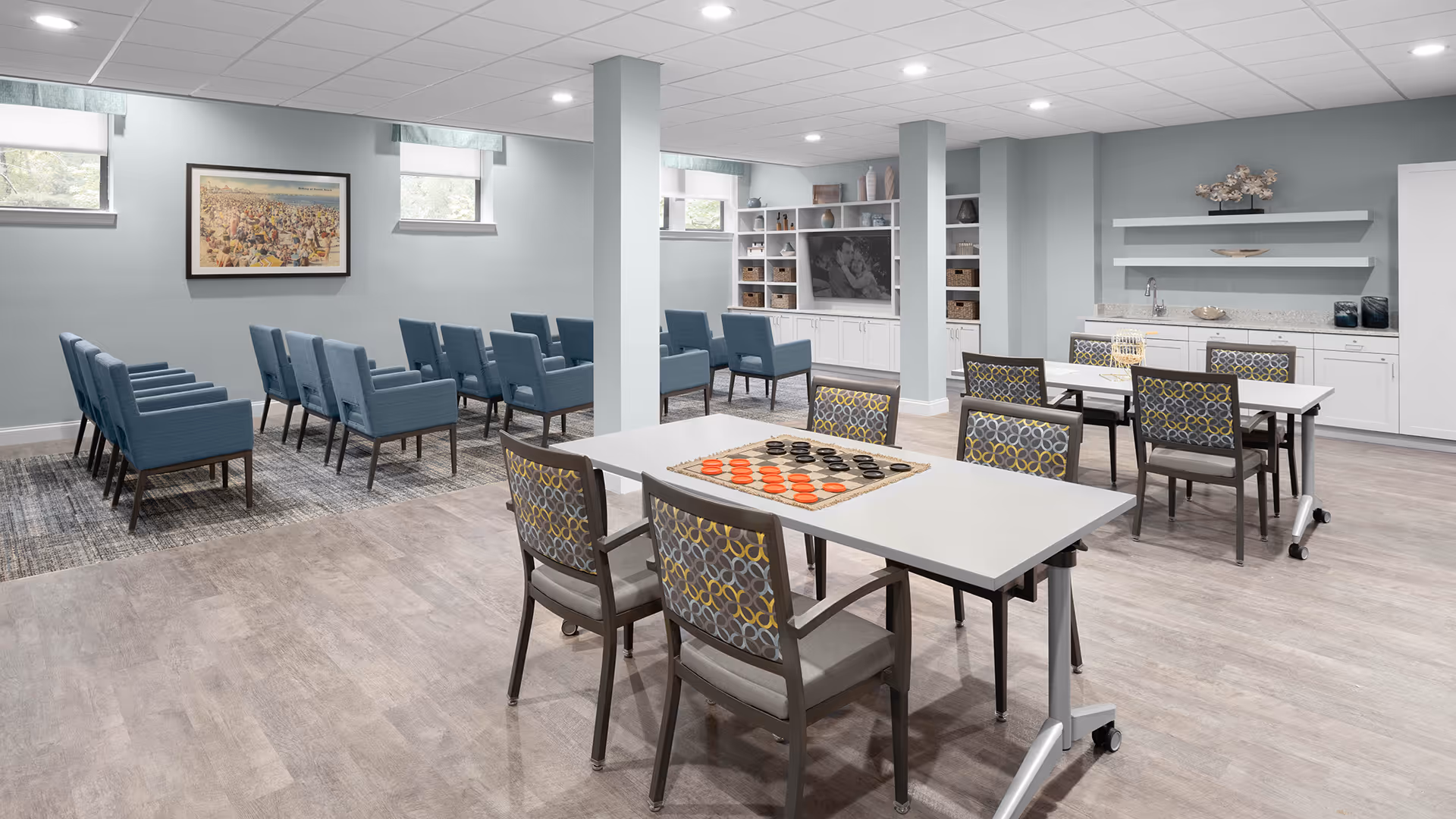 A spacious, well-lit common room with light blue walls and wood flooring. The room features two white tables with patterned chairs, one table has a checkers game set up. In the background, there are rows of blue upholstered chairs arranged facing a large TV mounted on a white built-in shelving unit. The room also has a countertop with a sink and decorative shelves above it.