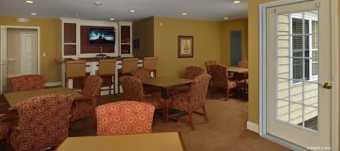 Community dining and lounge area with tables, patterned armchairs, a TV above a service counter, and a glass door to the exterior.