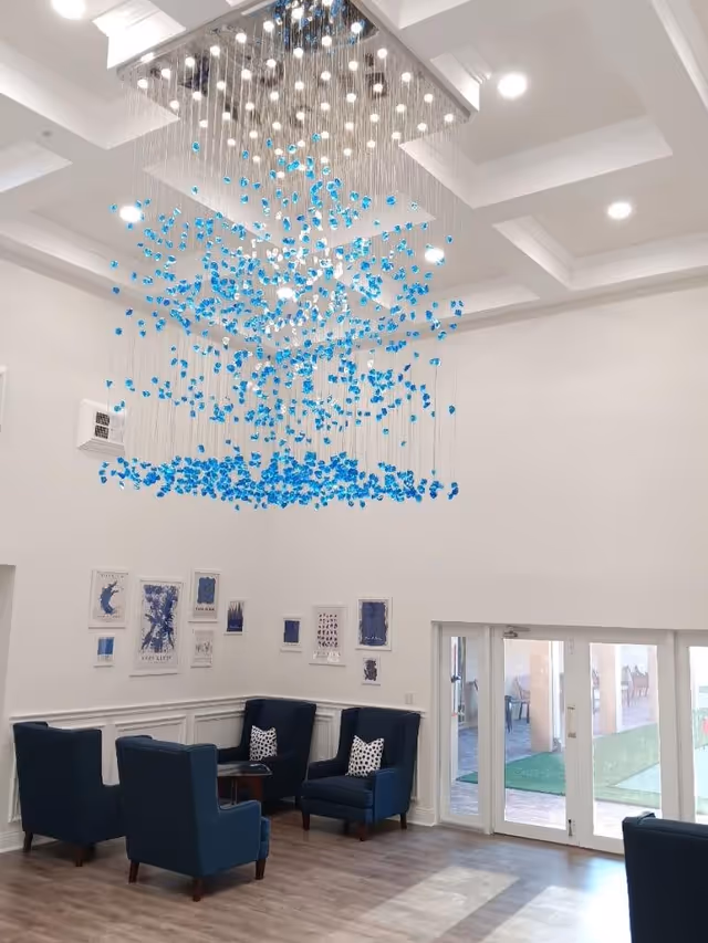 A bright and spacious common area with high ceilings featuring a modern chandelier with hanging blue and white decorative elements. The room has four dark blue armchairs arranged around a small wooden table. The walls are white with several framed artworks in blue tones. Large glass doors lead to an outdoor area with patio furniture and green artificial grass.
