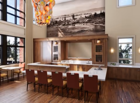 A spacious dining area with a large rectangular table surrounded by red chairs. The room features high ceilings with large windows allowing natural light to fill the space. A modern chandelier with orange and yellow glass elements hangs above the table. The back wall has a large black and white mural of a landscape and a built-in wooden cabinet with a mirror and sink. The floor is wooden, and there are additional tables and chairs near the windows.