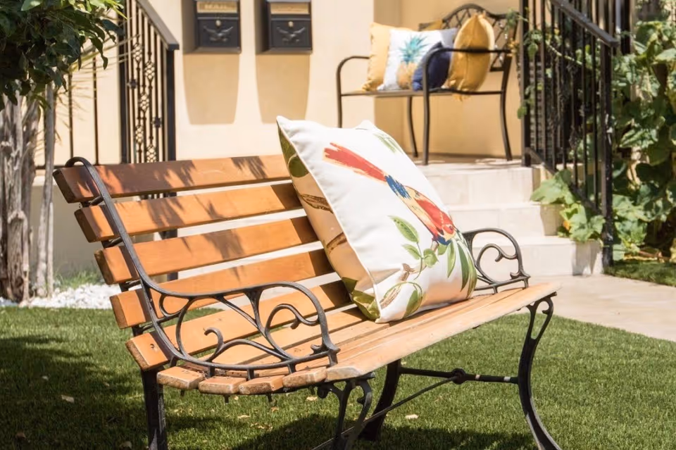 A wooden bench with black metal armrests and legs sits on a green lawn. The bench has a decorative pillow with a colorful bird design. In the background, there is a small staircase leading to a porch with another bench that has multiple pillows. The area is surrounded by plants and a beige building wall with two mailboxes.