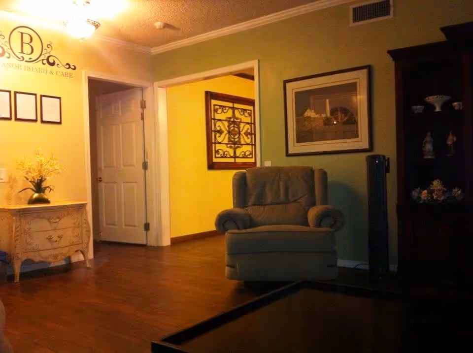 A cozy living room area with a comfortable armchair, a wooden cabinet displaying decorative items, a framed picture on the wall, and a small ornate table with a vase of yellow flowers. The room has wooden flooring and soft lighting, with an open doorway leading to another room with yellow walls.