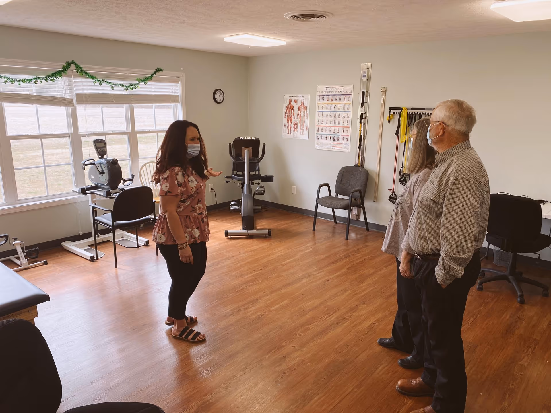 A woman wearing a mask and floral blouse is talking to an elderly man and woman, both also wearing masks, in a room with exercise equipment, chairs, and anatomical posters on the wall. The room has wooden flooring and large windows with blinds.