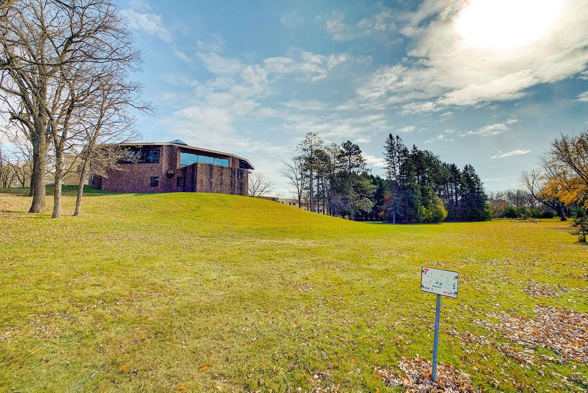 Grassy lawn and gently sloping hill with a brick building to the left, trees in the background, and a small sign in the foreground under a partly cloudy sky.