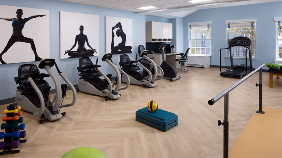 A senior living facility exercise room with four recumbent exercise bikes, a treadmill, a set of colorful dumbbells, a green exercise ball, a blue step platform with a yellow medicine ball on top, and a ballet barre. The walls are light blue with three large black and white silhouette yoga pose artworks. There are two windows letting in natural light and a small desk with a chair and cabinets in the corner.