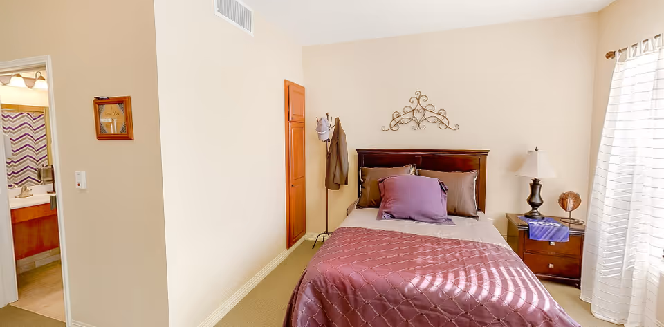 A bright bedroom with a bed dressed in purple bedding, a nightstand and lamp, a coat rack, and an open door showing an adjacent bathroom.