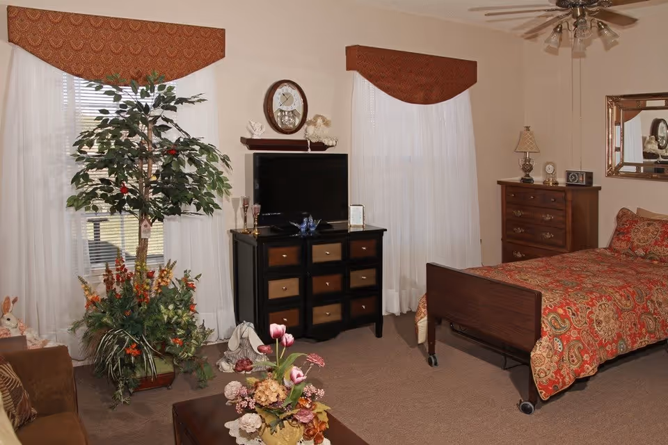 A cozy bedroom with a single bed covered in a red and gold patterned bedspread. The room features two large windows with white curtains and red valances. There is a black dresser with multiple drawers and a flat-screen TV on top, along with decorative items. A wooden chest of drawers with a lamp and clock sits against the wall. A ceiling fan with lights is mounted on the ceiling. A large potted plant and a coffee table with a floral arrangement add to the decor.