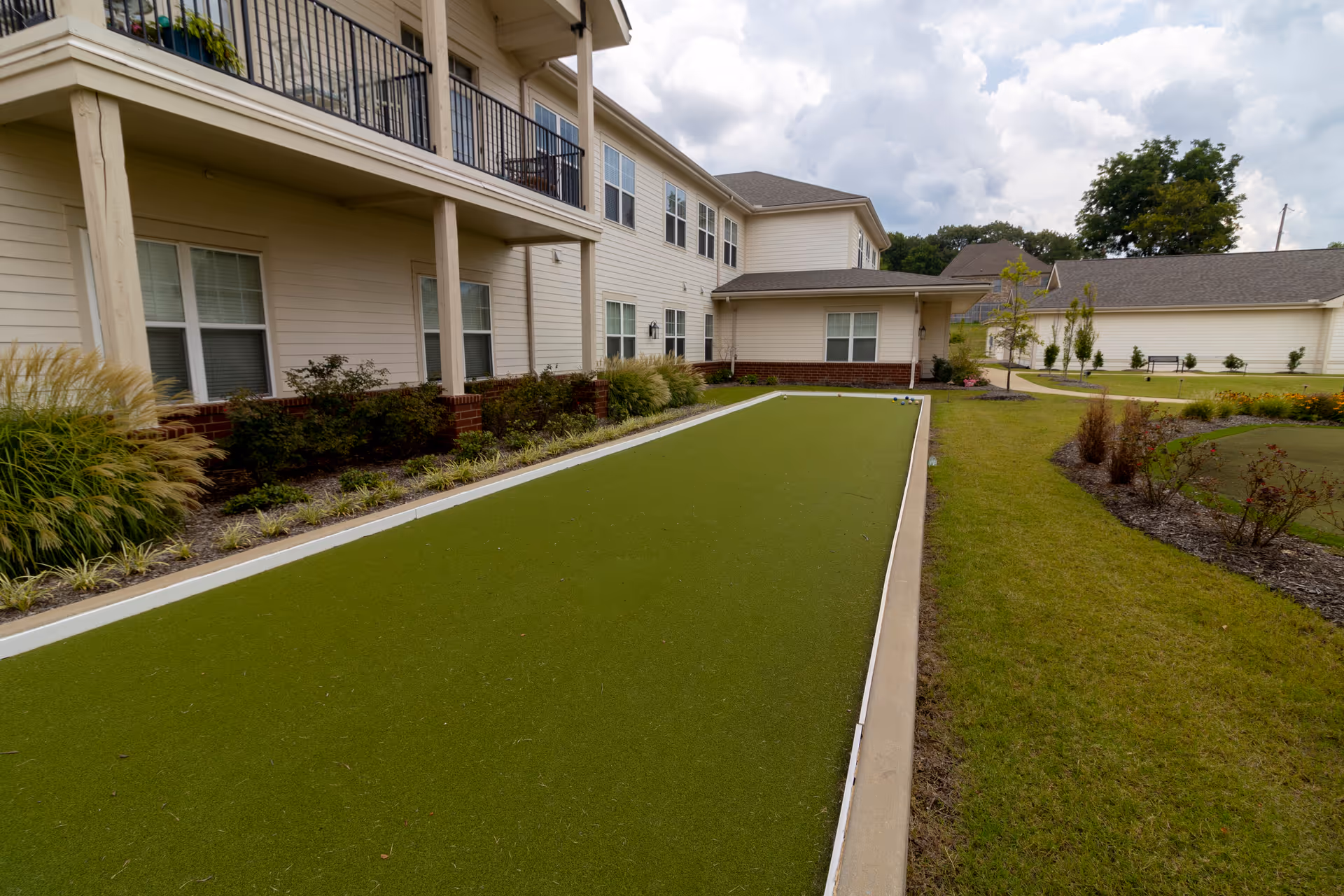 A bocce court with artificial turf beside a two-story senior living building and landscaped grounds.