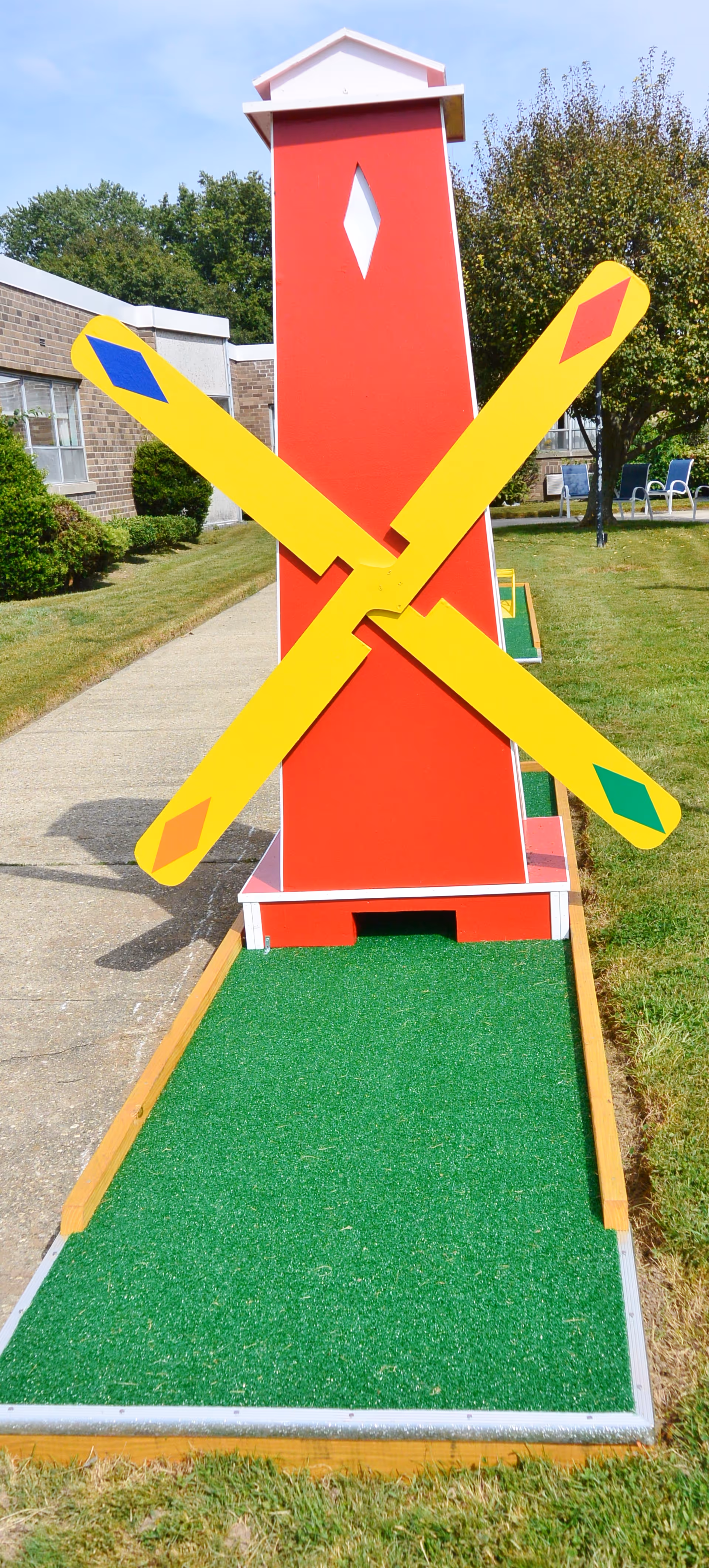 A brightly colored miniature golf course obstacle shaped like a red windmill with yellow blades featuring colored diamond shapes. The windmill is situated on a green artificial turf putting surface with a sidewalk and building visible in the background, along with trees and outdoor chairs.