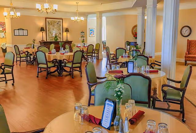 A bright and spacious dining room in a senior living facility with round tables covered in beige tablecloths, each set with glasses, napkins, and small flower vases. The room features wooden flooring, green cushioned chairs, chandeliers hanging from the ceiling, and decorative elements such as framed artwork and lamps on a sideboard.