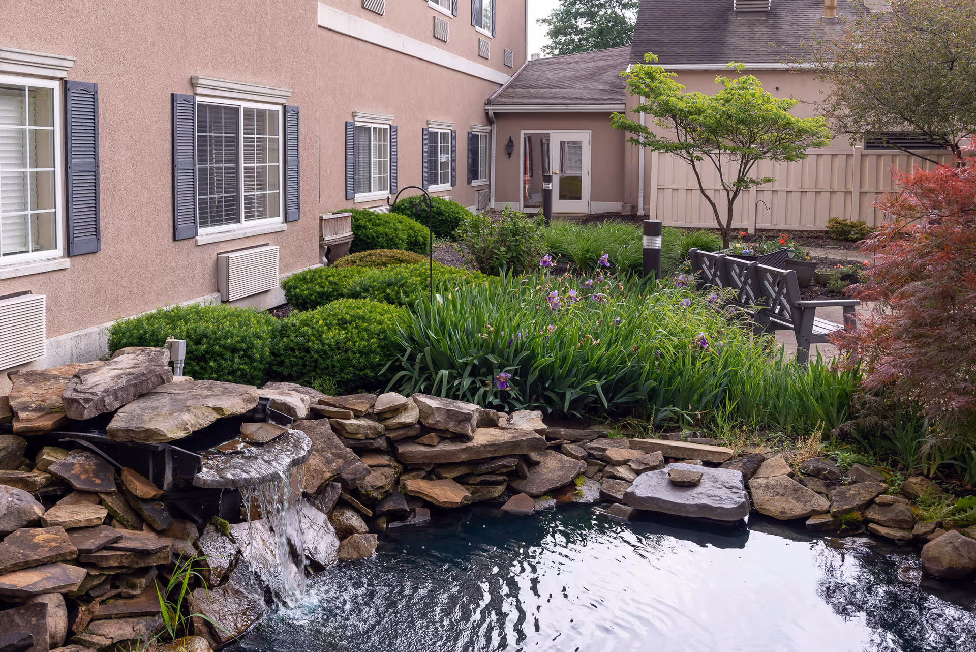 Outdoor garden area at The Abbewood featuring a small pond with a waterfall, surrounded by rocks, green shrubs, flowering plants, and trees. There is a bench near the pond and a beige building with windows and shutters in the background.