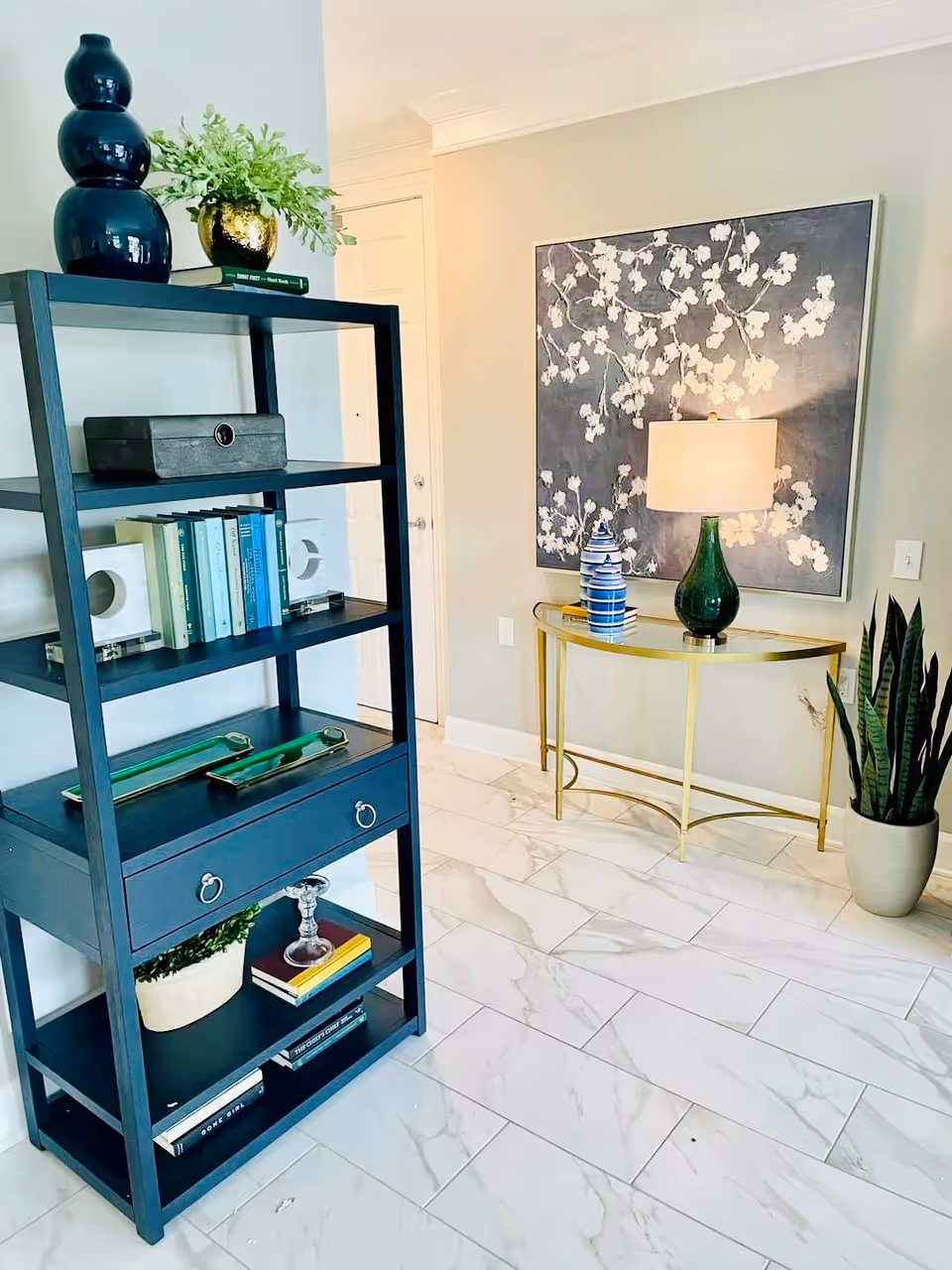 A modern interior corner featuring a dark blue shelving unit with books, decorative items, and plants. Next to it is a gold console table with a green lamp, blue and white decorative jars, and a large floral painting on the wall. The floor is tiled with white marble-like tiles, and there is a tall potted plant on the right side.