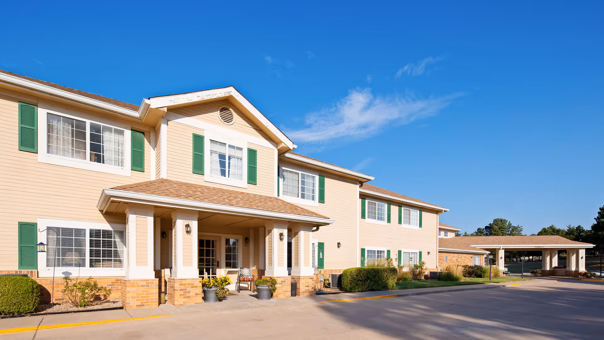 Exterior view of a two-story senior living facility building with beige siding, green window shutters, and a covered entrance with columns. There are potted plants and chairs near the entrance, and a clear blue sky overhead.