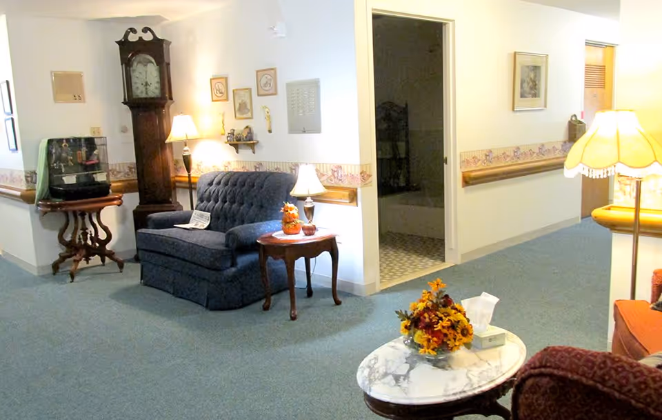 Interior of a senior living facility hallway with a blue upholstered loveseat, a wooden grandfather clock, side tables with lamps, a small fish tank on a wooden table, and a round marble-top table with a flower arrangement and tissue box. The walls have a decorative border and handrails, and there is an open doorway leading to a tiled room.
