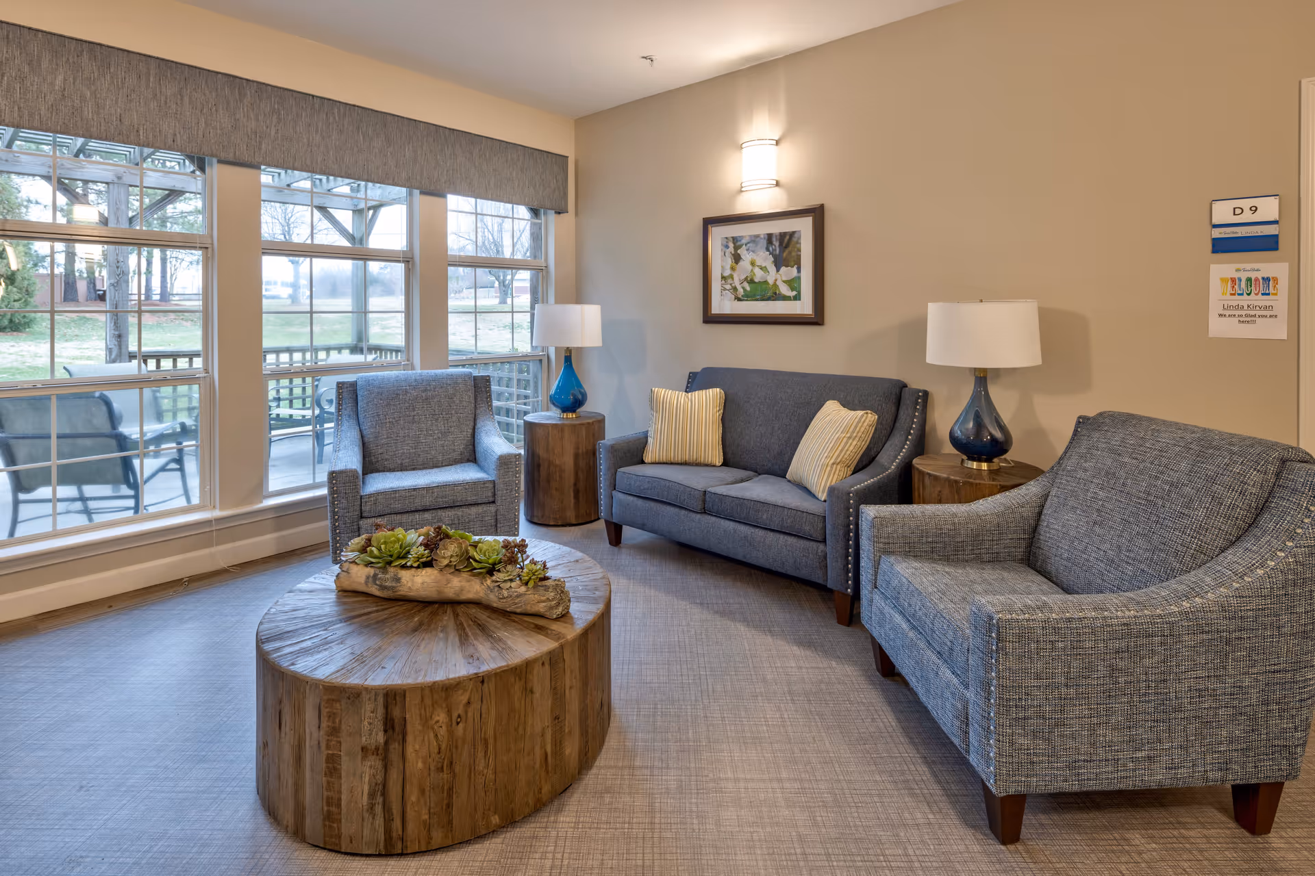 Cozy seating area with two armchairs and a loveseat around a round wooden coffee table beside large windows overlooking a patio.