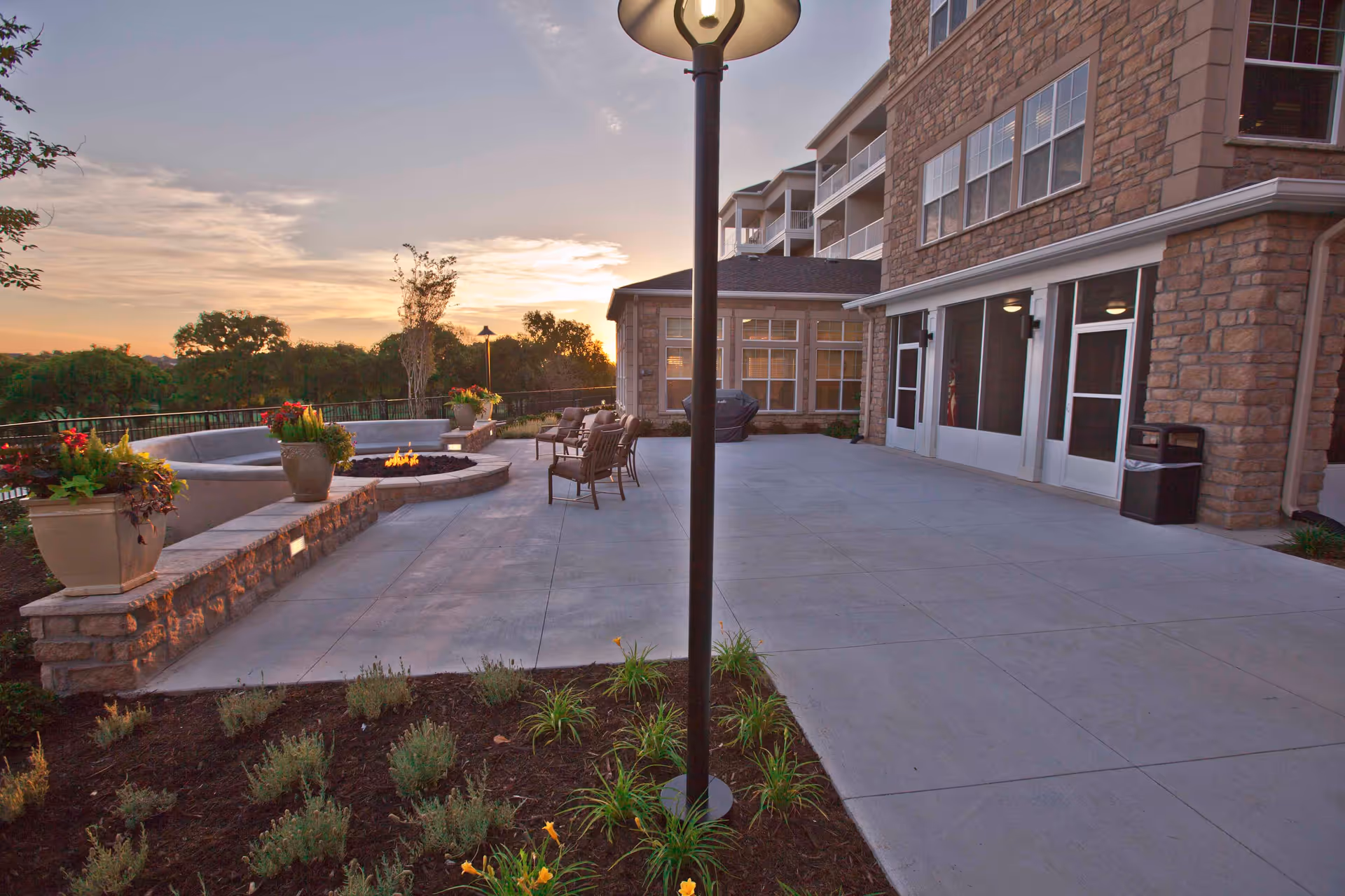 Outdoor patio at a senior living facility with a stone building, seating around a fire pit, potted plants, and a lamppost at sunset.