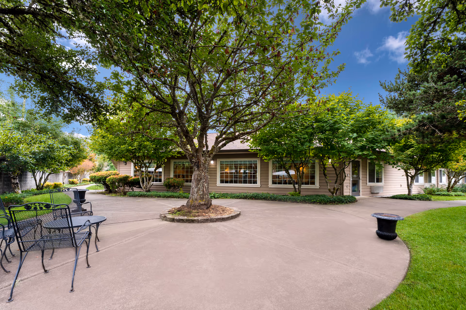 Outdoor courtyard area at The Hampton featuring a large tree in the center of a paved circular walkway, surrounded by green grass, bushes, and other trees. There are black metal chairs and a table on the left side, and a building with large windows and a door in the background under a partly cloudy blue sky.