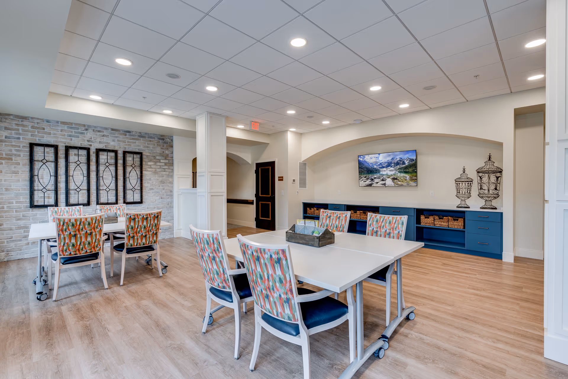 A bright and spacious common area with wooden flooring, featuring two white tables with colorful patterned chairs around them. The room has a modern ceiling with recessed lighting, a brick accent wall with decorative framed panels, and a blue built-in cabinet with baskets and a flat-screen TV mounted above it displaying a mountain landscape. The space appears clean and inviting with neutral walls and an arched alcove.