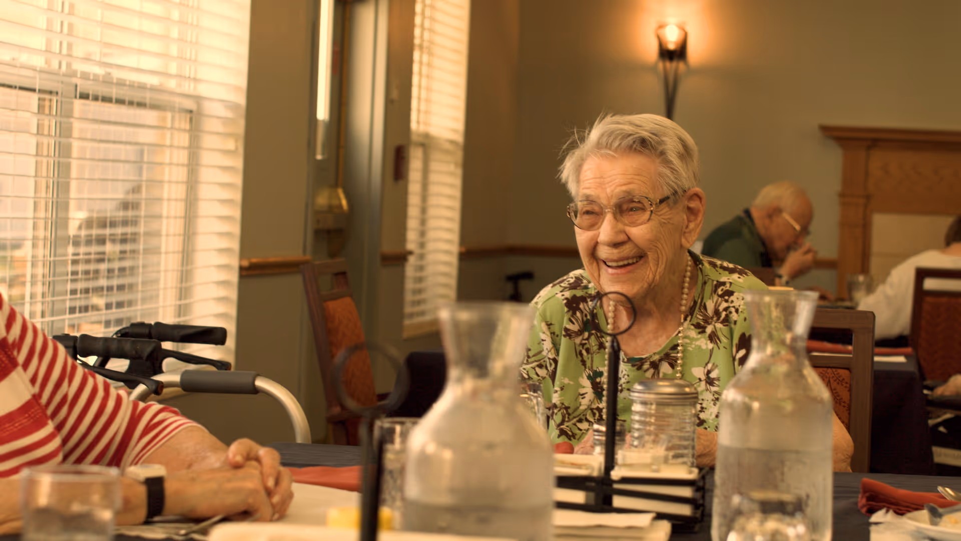 An elderly woman with short gray hair and glasses, wearing a green floral blouse, smiling and sitting at a dining table in a well-lit room with large windows and blinds. There are water pitchers, glasses, and condiments on the table. Other elderly people are seated in the background.