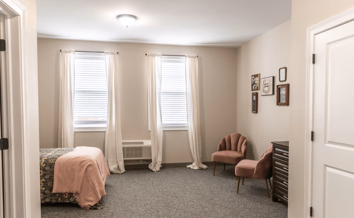 A cozy bedroom with two windows covered by white curtains, a bed with a floral bedspread and a pink blanket, two pink upholstered chairs, a dark wooden dresser, and framed artwork on the wall.