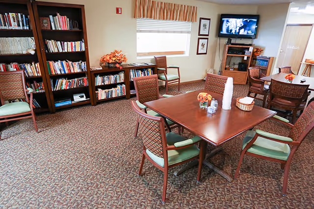 A senior living common room with wooden tables and chairs, bookshelves, a television, and seasonal decorations.