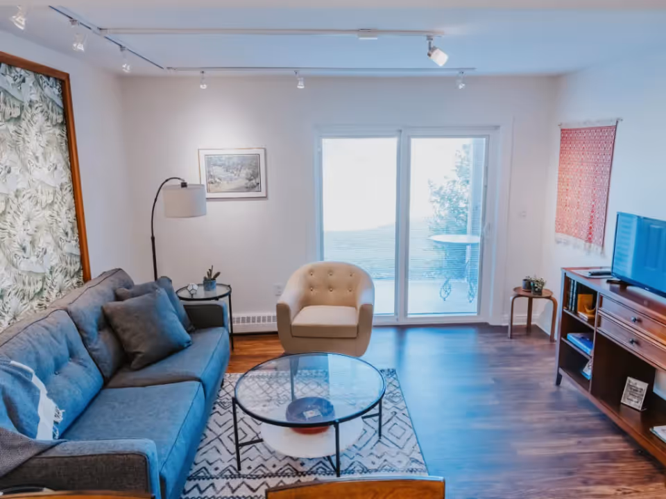 Bright living room with a gray sofa, beige armchair, glass coffee table, TV console and sliding glass doors to the outside.