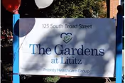 Outdoor sign for The Gardens at Lititz located at 125 South Broad Street, with Priority Healthcare Group mentioned below. The sign is white with blue and green text, surrounded by greenery and balloons.