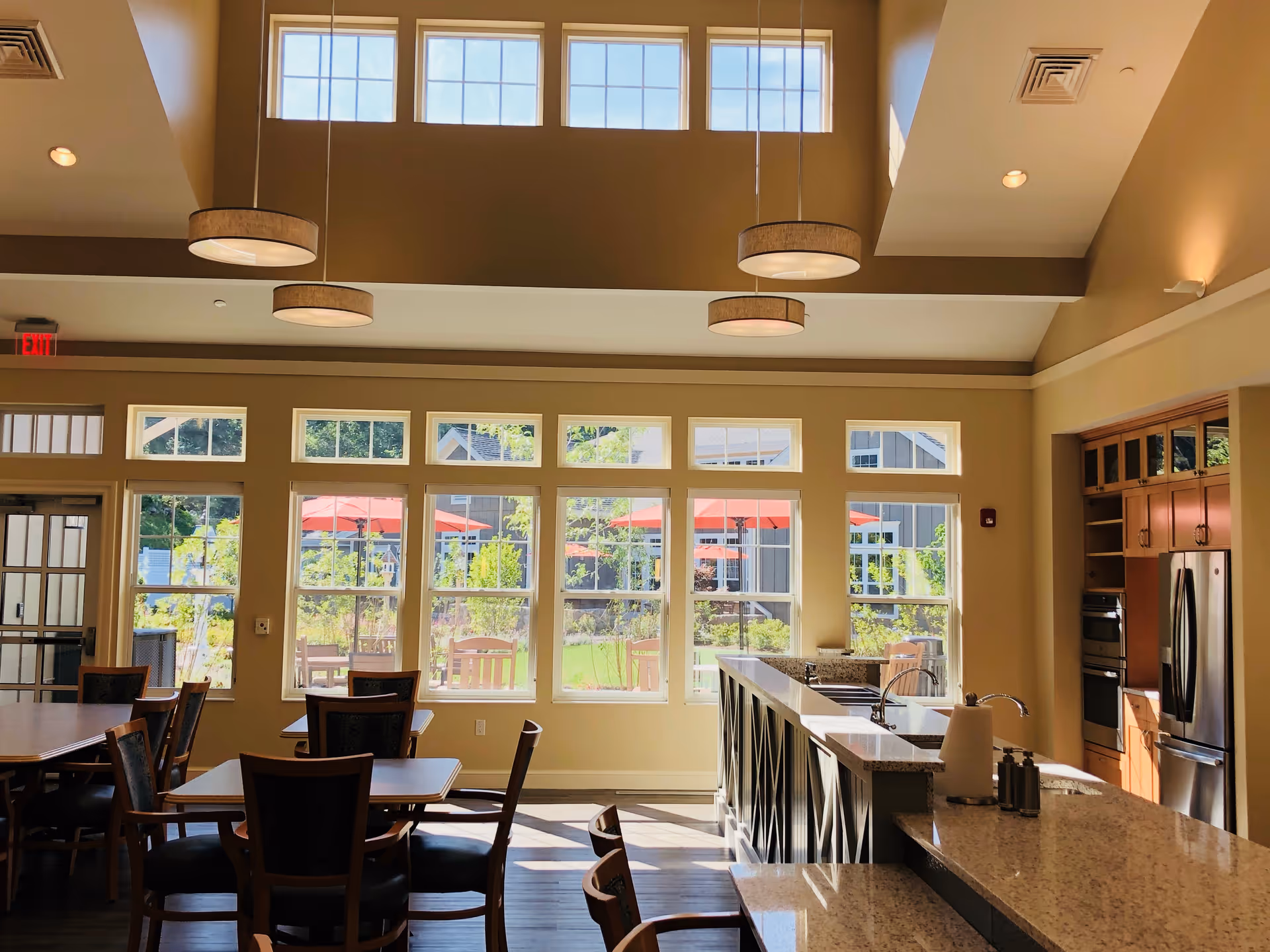 Bright and spacious dining area with multiple wooden tables and chairs, large windows letting in natural light, and a kitchen area with granite countertops, stainless steel appliances, and pendant lights hanging from a high ceiling.