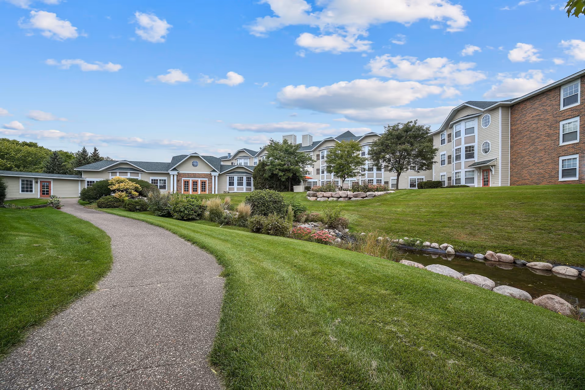 A paved walkway curves through a well-maintained grassy area with shrubs and flowers, leading to a large multi-story building with beige siding and brick accents under a partly cloudy blue sky.
