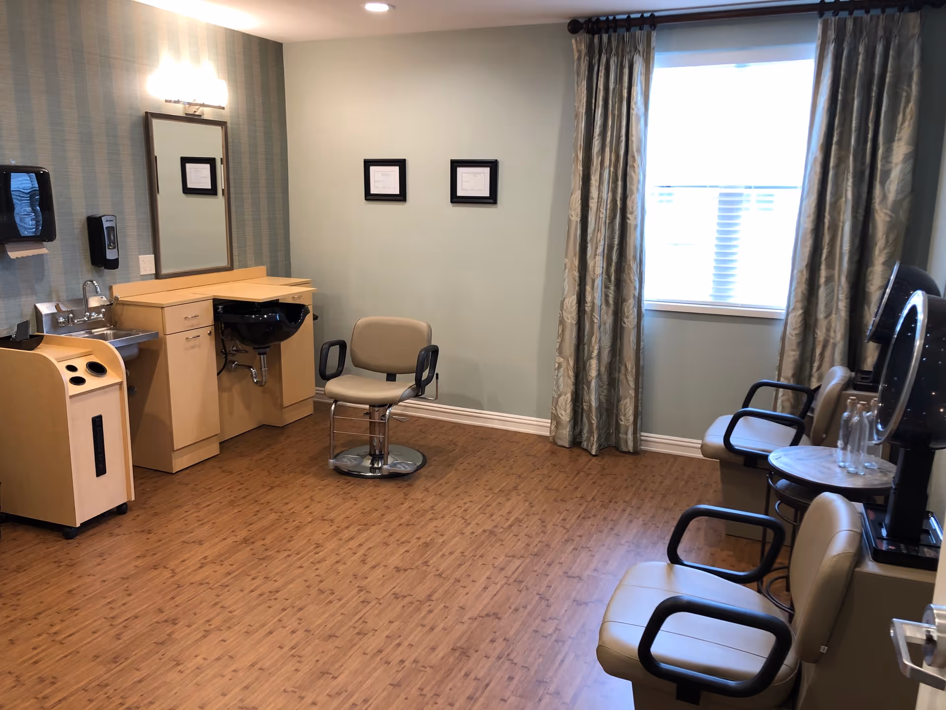 Interior of a hair salon room with wooden flooring, a styling chair in front of a mirror and sink, two waiting chairs with armrests, a hair dryer, and a window with patterned curtains.