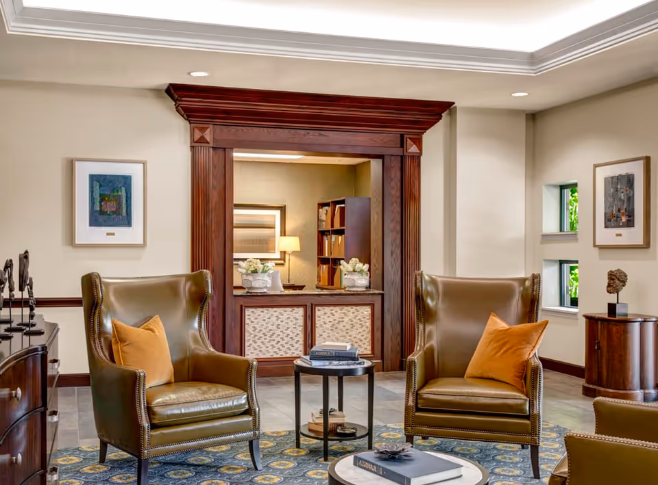 A cozy sitting area with two brown leather armchairs, each with an orange throw pillow, positioned around a small round table with books on it. The room features a decorative wooden frame around an open window to another room with a lamp and bookshelf. The walls are adorned with framed artwork, and there is a patterned rug on the floor.