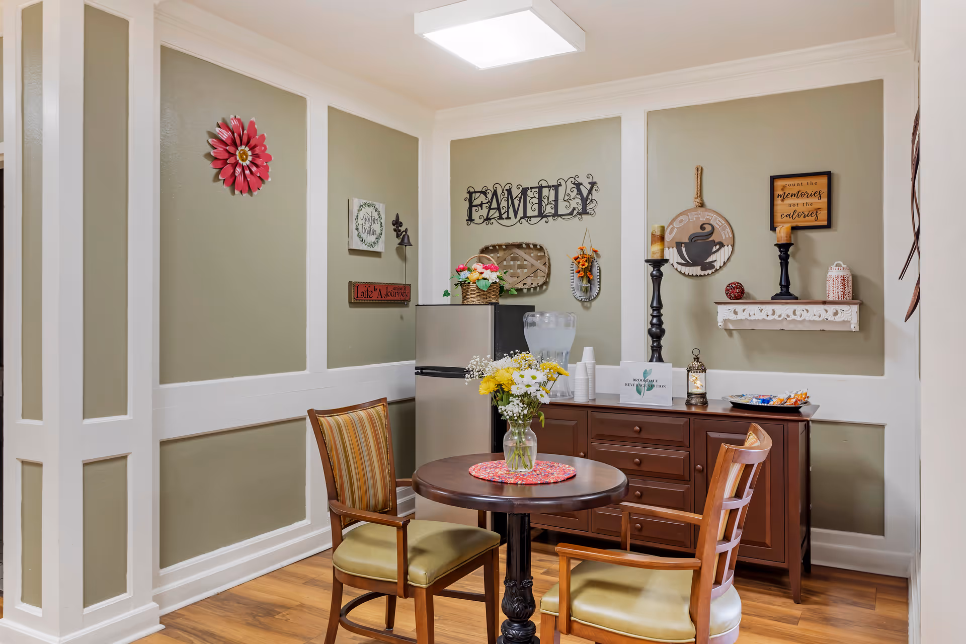 A cozy corner in a senior living facility with a small round wooden table and two cushioned chairs. On the table is a vase with fresh flowers. Behind the table is a wooden cabinet with a water dispenser, cups, and a small refrigerator. The walls are decorated with various wall art including the word 'FAMILY', a coffee sign, and inspirational quotes. The room has light green walls with white trim and wooden flooring.