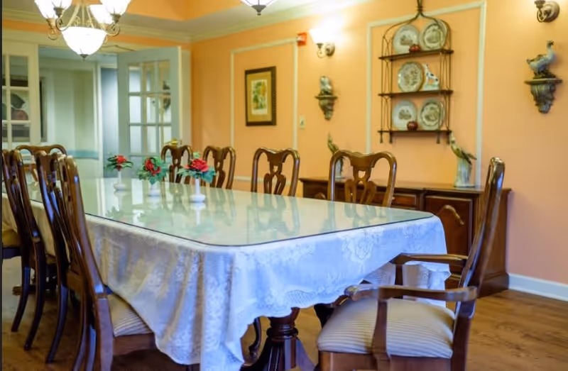 A formal dining room with a long rectangular table covered with a white lace tablecloth and a glass top. The table is surrounded by wooden chairs with cushioned seats. On the table are small vases with red flowers. The walls are painted peach and decorated with framed artwork, wall sconces, and a decorative plate rack. The floor is wooden, and a chandelier hangs from the ceiling.