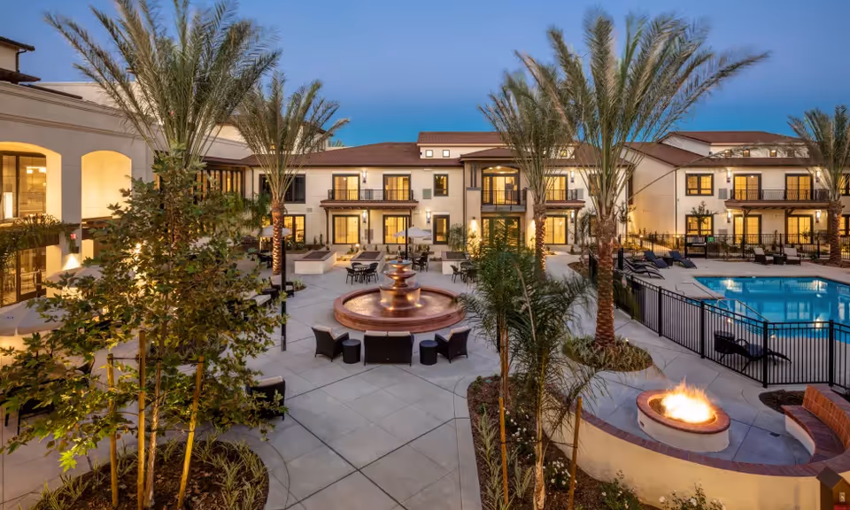 Outdoor courtyard area of Merrill Gardens at Rancho Cucamonga featuring a central water fountain surrounded by seating, palm trees, a swimming pool with lounge chairs, and a fire pit with curved seating. The building with balconies and large windows is visible in the background during dusk.