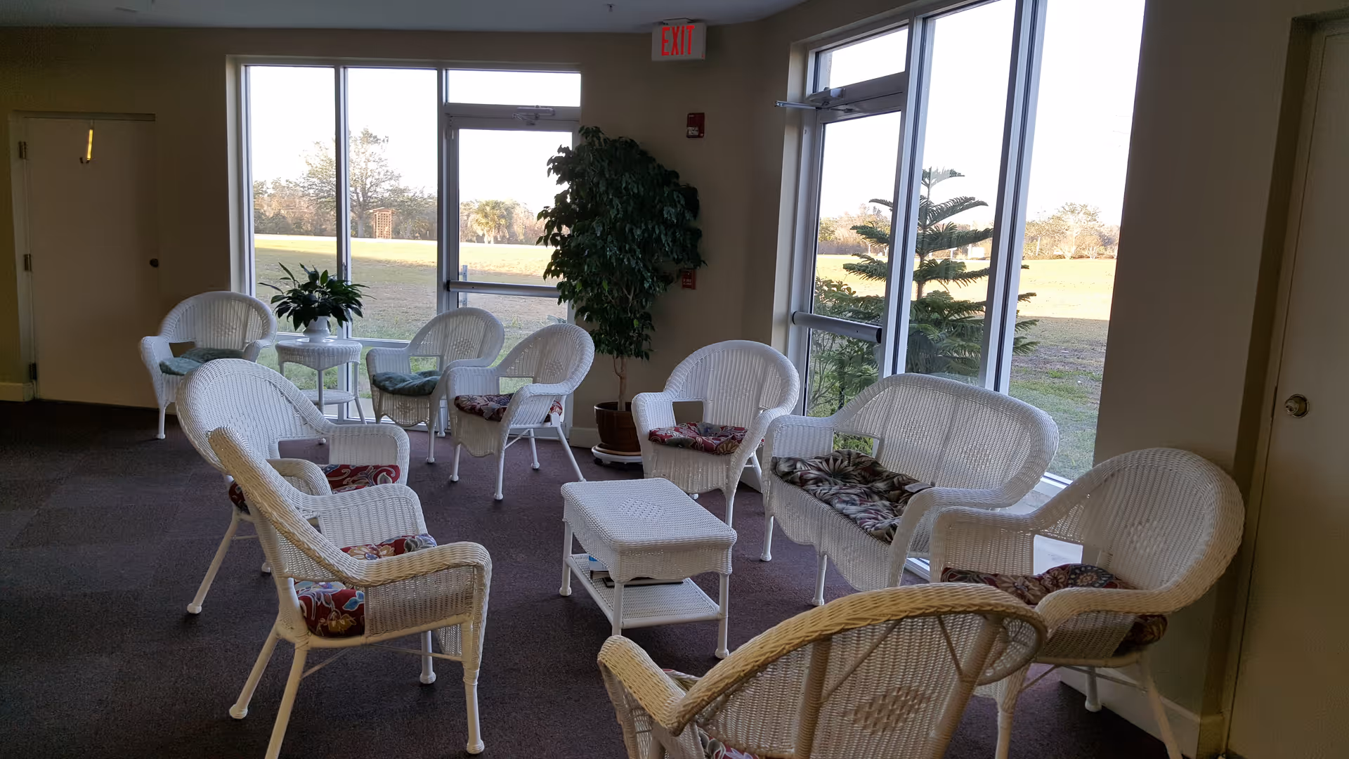 A bright common area with white wicker chairs and a wicker loveseat arranged around a small white wicker coffee table. The chairs have colorful cushions, and there are large windows and glass doors letting in natural light and showing an outdoor grassy area. A potted plant is placed near the windows.