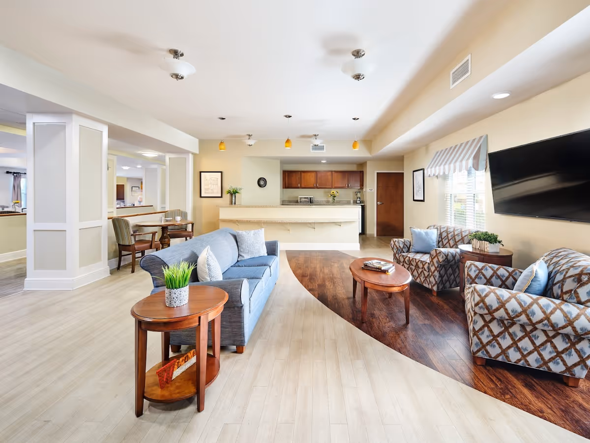 A bright and spacious living room area in a senior living facility with a blue sofa, two patterned armchairs with blue cushions, a wooden coffee table, and a side table with a small plant. The room features a large flat-screen TV mounted on the wall, a window with striped valance, and a kitchen area with wooden cabinets and a countertop in the background. There is also a small dining table with chairs near a white column.
