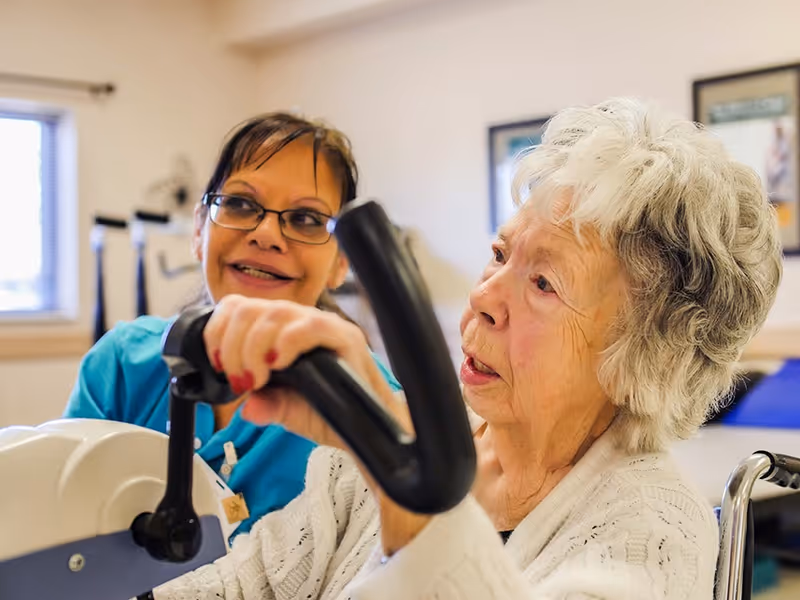 An elderly woman using a hand cycle exercise machine with the assistance of a smiling caregiver in a rehabilitation or healthcare center room.