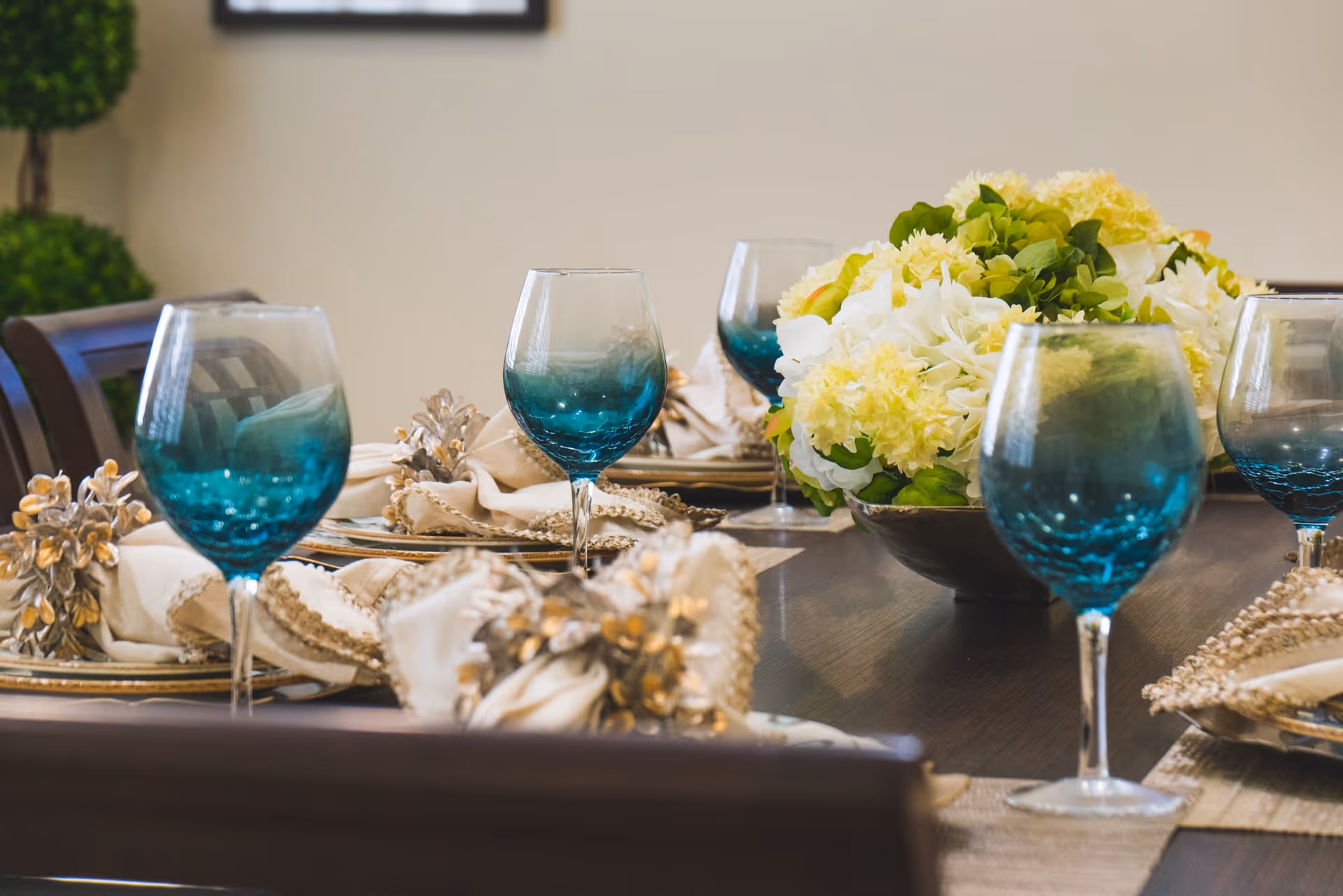 A close-up view of a dining table set with blue-tinted wine glasses, beige napkins with decorative napkin rings, and a centerpiece of white and yellow flowers with green leaves.