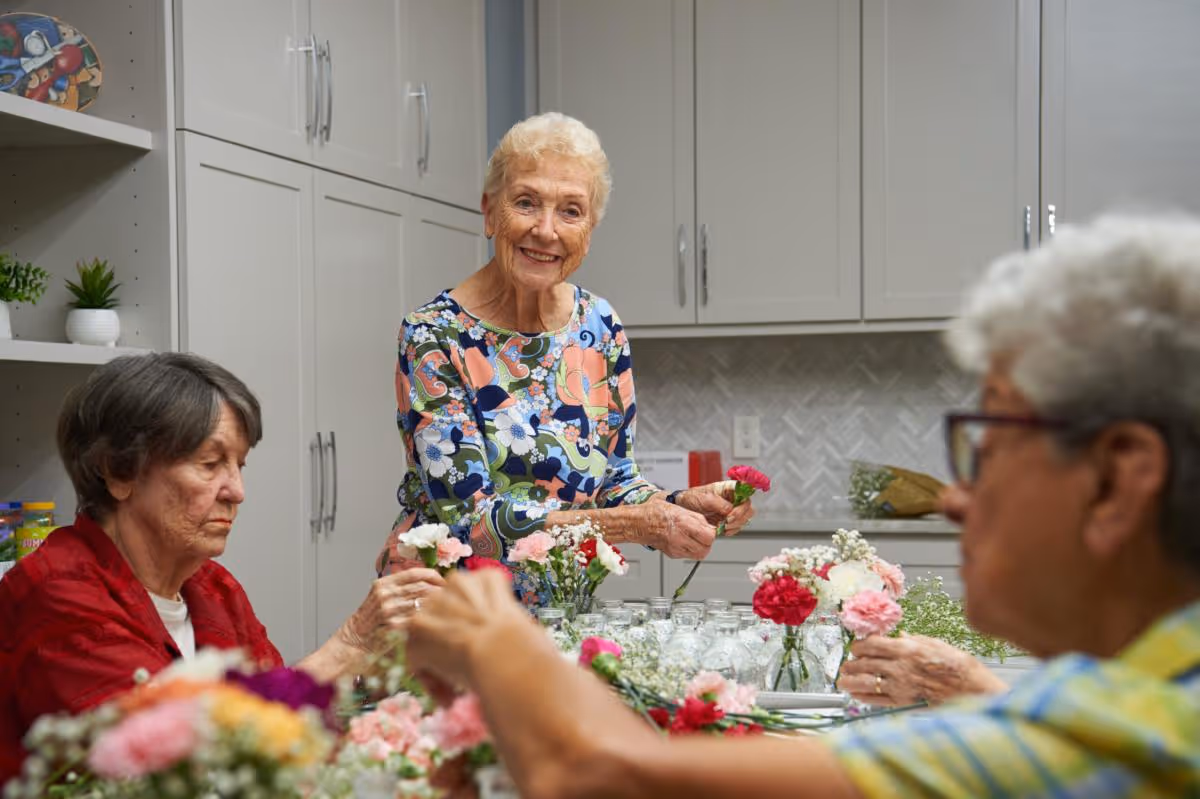 Three elderly women arranging flowers in small glass jars on a table in a kitchen area with white cabinets and a herringbone tile backsplash. One woman in a colorful floral top is smiling at the camera while holding a pink carnation.
