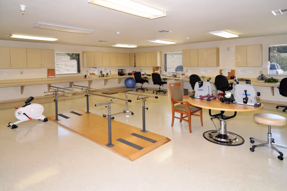 A spacious rehabilitation or physical therapy room with parallel bars for walking exercises, stationary exercise bikes, a large exercise ball, and various chairs and desks along the walls. The room has beige cabinets, multiple windows with blinds, and bright overhead lighting.