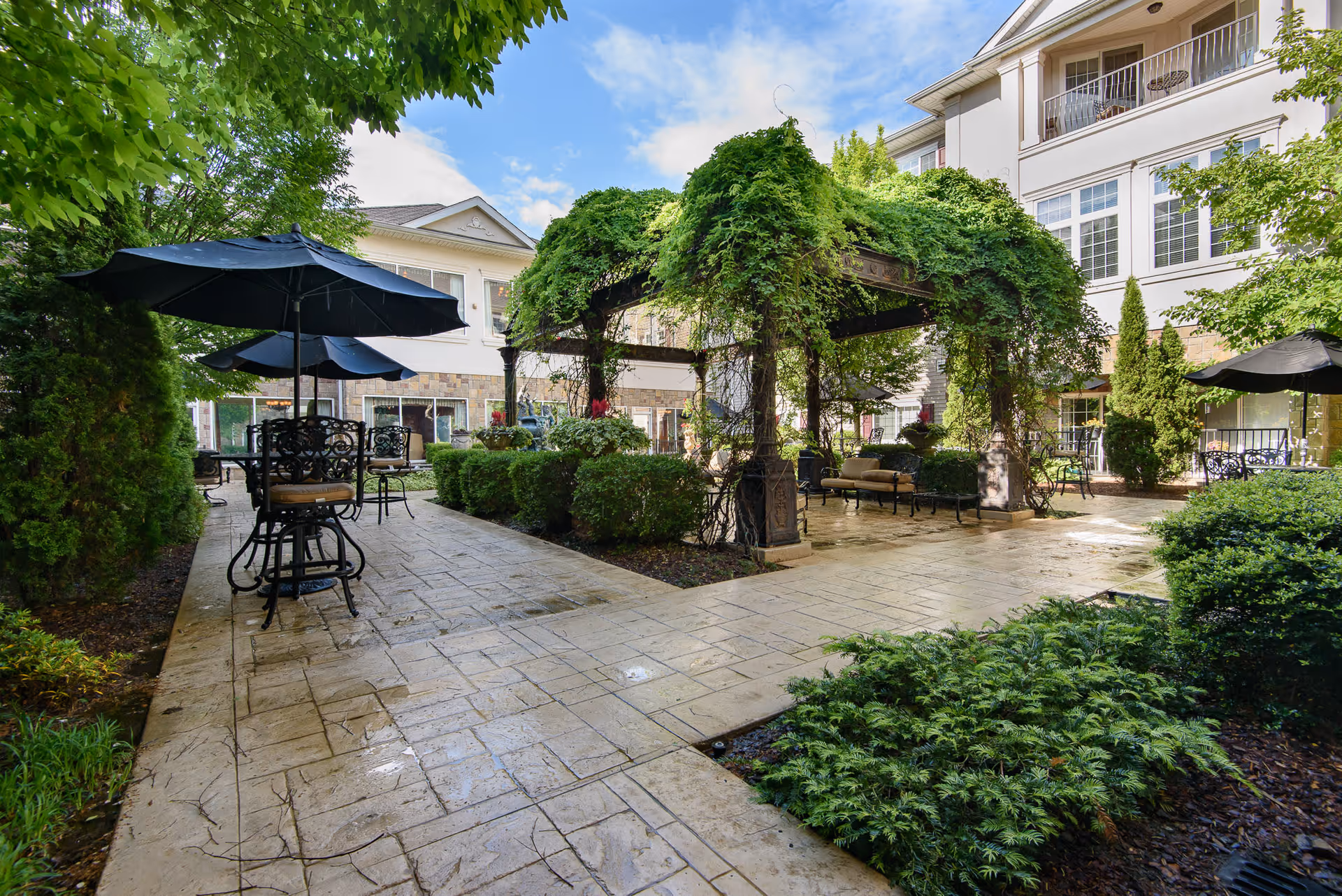 Courtyard with paved walkways, umbrella-shaded tables and a vine-covered pergola in front of the facility building.
