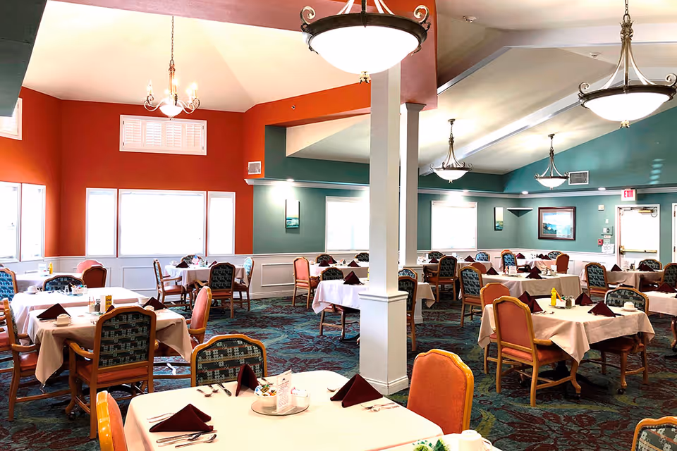 A spacious dining room with multiple tables covered in white tablecloths, each set with silverware, napkins, and condiments. The room features colorful walls painted in red and teal, patterned carpet, and several chandeliers hanging from the ceiling providing warm lighting. Chairs around the tables have a mix of orange and patterned upholstery. Large windows allow natural light to brighten the space.