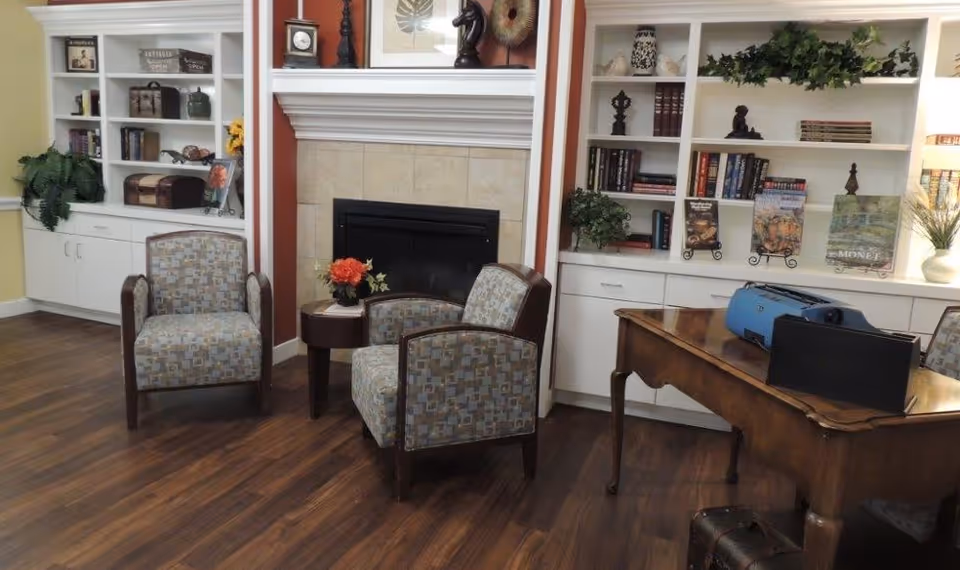 A cozy communal sitting area with a fireplace flanked by built-in bookshelves, three upholstered chairs, a small side table with flowers, and a wooden desk.